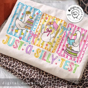 May include: A white t-shirt with a colourful graphic of three cartoon ducks, books, and the text "JUST A SILLY TEST." Each duck is in a different frame with a unique design. The shirt is folded on a leopard print fabric.