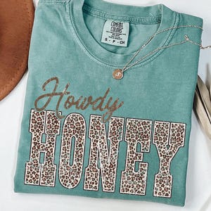 May include: A teal Comfort Colors t-shirt with the words "Howdy HONEY" in a brown script and leopard print block letters. A gold necklace with a pendant is draped on the shirt.
