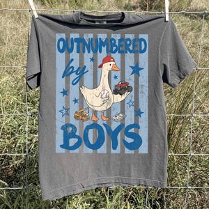 May include: Gray t-shirt with a graphic featuring a goose wearing a baseball cap, holding a baseball and a monster truck. The text reads "OUTNUMBERED by BOYS" in blue. Stars and stripes complete the design.