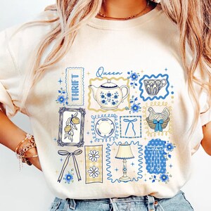 May include: Cream-colored sweatshirt featuring a collage of vintage-style illustrations in blue and yellow. Designs include a teapot, teacup, lemons, a chicken in a basket, and the word "Thrift". The word "Queen" is also present.