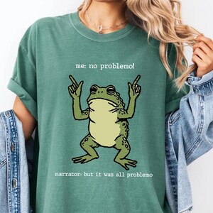 May include: Green t-shirt featuring a cartoon frog with the text "me: no problemo!" above it and "narrator: but it was all problemo" below. The frog is green with a light green belly and is raising its hands.