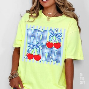 May include: A neon yellow t-shirt with the word "MOM" in blue gingham letters, adorned with red cherries, white flowers, and blue bows. The shirt has a relaxed fit and short sleeves.
