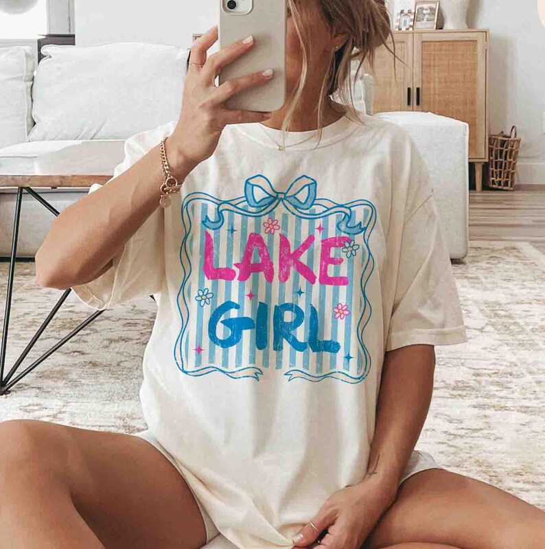 May include: Cream-coloured t-shirt featuring the words "LAKE GIRL" in pink and blue, framed by a blue border with a bow and floral accents. The shirt has a striped background and is a casual, oversized style.