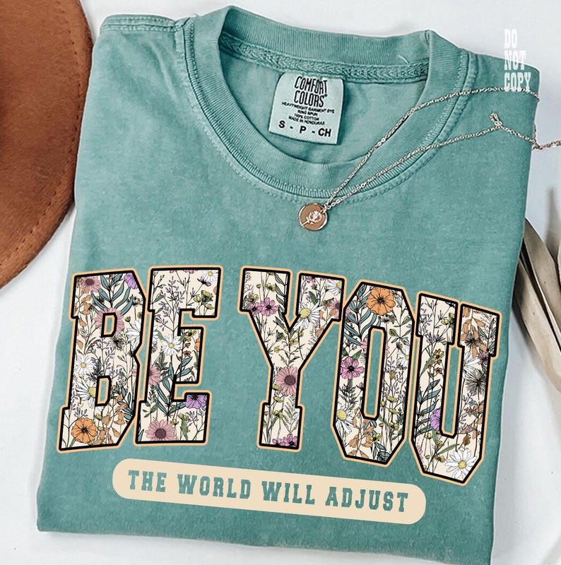 May include: A teal Comfort Colors t-shirt with the words "BE YOU" in a floral design. Below the text is the phrase "THE WORLD WILL ADJUST". A silver necklace with a pendant rests on the shirt.