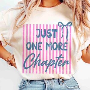 Puede incluir: Camiseta color crema con el texto azul "JUST ONE MORE CHAPTER". El texto est&aacute; sobre un fondo de rayas rosas y blancas, con un lazo azul. La camiseta es de estilo casual.