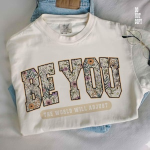 May include: Cream-colored t-shirt with the words "BE YOU" in a floral design. Below the text is the phrase "THE WORLD WILL ADJUST" in a rectangular banner. The shirt is folded on top of blue jeans.