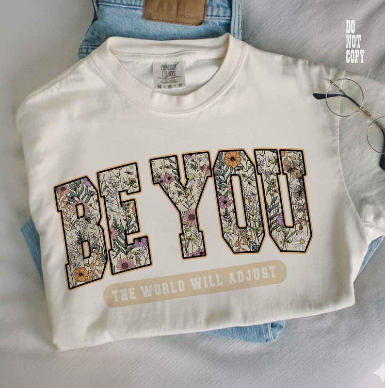 May include: Cream-colored t-shirt with the words "BE YOU" in a floral design. Below the text is the phrase "THE WORLD WILL ADJUST" in a rectangular banner. The shirt is folded on top of blue jeans.