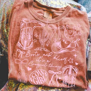 May include: A brown t-shirt with a pink graphic design featuring cowboy boots, a cactus, a horseshoe, a cowboy hat, and the words "Boots and bling it's a Cowgirl thing" and "Cow Girl".