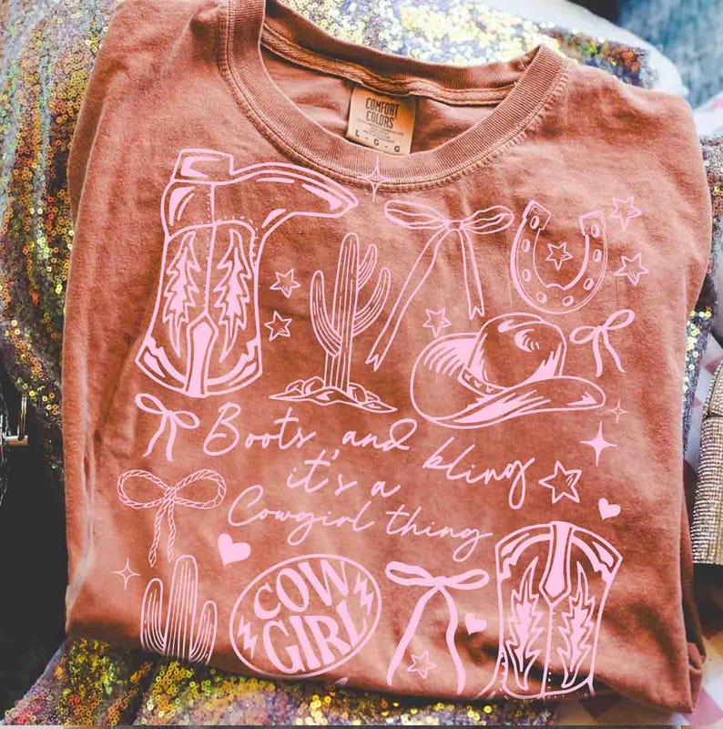 May include: A brown t-shirt with a pink graphic design featuring cowboy boots, a cactus, a horseshoe, a cowboy hat, and the words "Boots and bling it's a Cowgirl thing" and "Cow Girl".