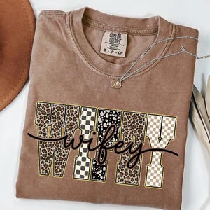 May include: A brown Comfort Colors t-shirt featuring the word "wifey" in a decorative font. The letters are patterned with leopard print, black and white checkered, and polka dot designs. A silver necklace with a pendant is draped across the shirt.