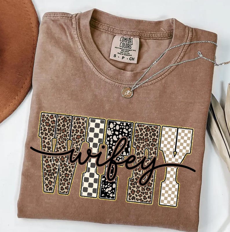 May include: A brown Comfort Colors t-shirt featuring the word "wifey" in a decorative font. The letters are patterned with leopard print, black and white checkered, and polka dot designs. A silver necklace with a pendant is draped across the shirt.