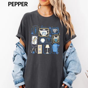 May include: Dark gray t-shirt with a collage of blue and white illustrations, including a teapot, teacup, and floral designs. The word "THRIFT" is vertically printed on the left side of the shirt, and the word "Queen" is printed above the teapot.
