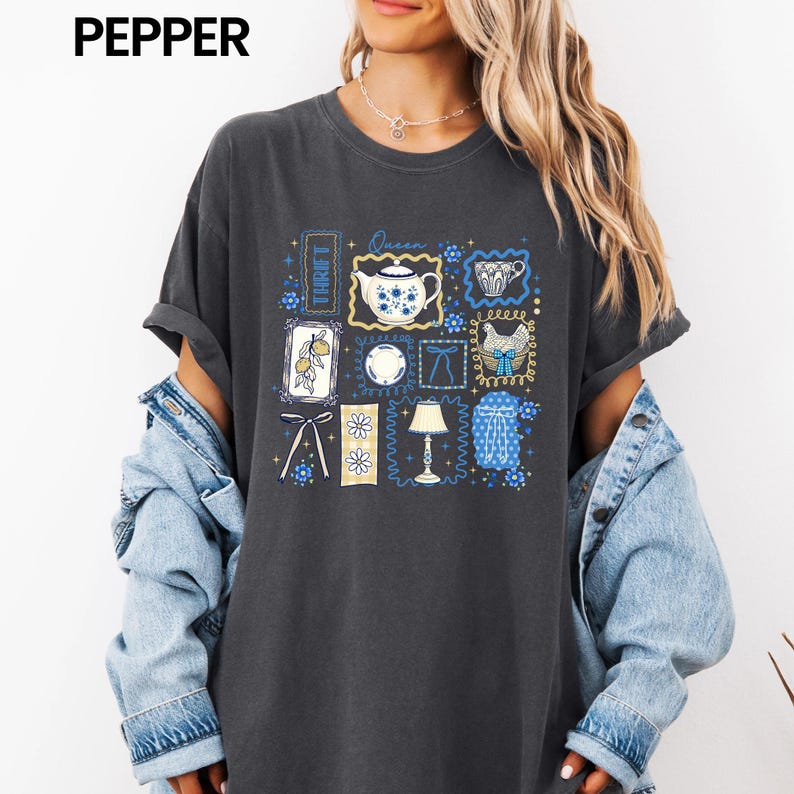 May include: Dark gray t-shirt with a collage of blue and white illustrations, including a teapot, teacup, and floral designs. The word "THRIFT" is vertically printed on the left side of the shirt, and the word "Queen" is printed above the teapot.