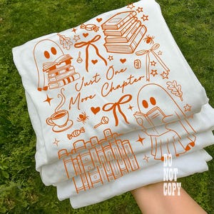 May include: White t-shirts with orange illustrations of books, ghosts, and the text "Just One More Chapter." The design includes bows, stars, and a coffee cup. The shirts are stacked, with the top one slightly visible.