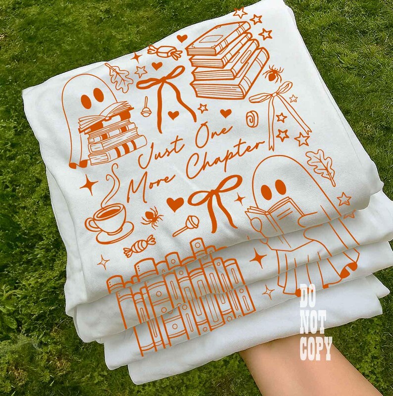 May include: White t-shirts with orange illustrations of books, ghosts, and the text "Just One More Chapter." The design includes bows, stars, and a coffee cup. The shirts are stacked, with the top one slightly visible.