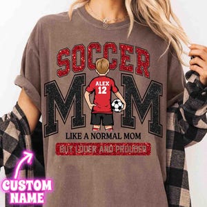 May include: Brown t-shirt with the words "SOCCER MOM" in red glitter and black letters. A graphic of a soccer player in a red jersey with the name "ALEX" and the number "12" is in the center. The text "LIKE A NORMAL MOM BUT LOUDER AND PROUDER" is below.