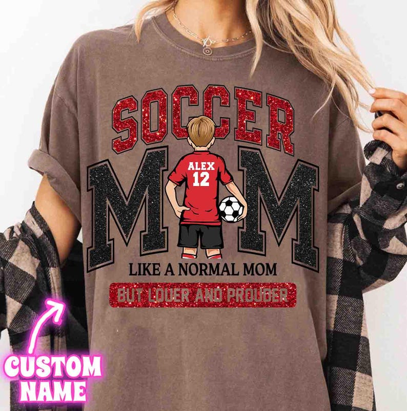 May include: Brown t-shirt with the words "SOCCER MOM" in red glitter and black letters. A graphic of a soccer player in a red jersey with the name "ALEX" and the number "12" is in the center. The text "LIKE A NORMAL MOM BUT LOUDER AND PROUDER" is below.