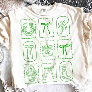 May include: Cream-colored t-shirt with a green St. Patrick's Day design. The design features a horseshoe, bows, a four-leaf clover, a pot of gold, and coins. The shirt is made of a soft material.