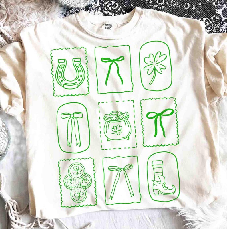 May include: Cream-colored t-shirt with a green St. Patrick's Day design. The design features a horseshoe, bows, a four-leaf clover, a pot of gold, and coins. The shirt is made of a soft material.