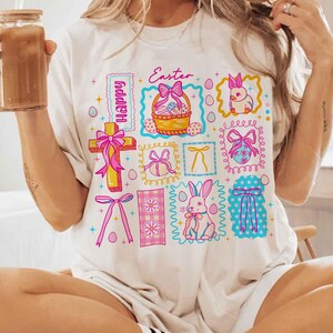 May include: Cream-colored t-shirt featuring a colorful Easter-themed graphic. The design includes a cross, a basket of eggs, a bunny, and the word "Easter". Pink and blue accents are used throughout the design.