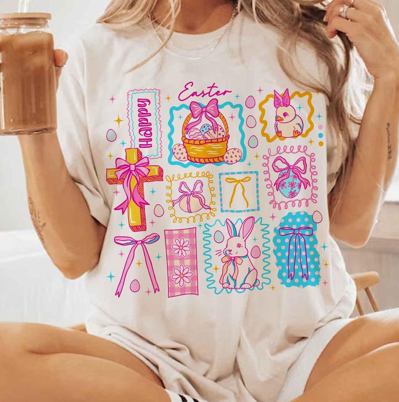 May include: Cream-colored t-shirt featuring a colorful Easter-themed graphic. The design includes a cross, a basket of eggs, a bunny, and the word "Easter". Pink and blue accents are used throughout the design.