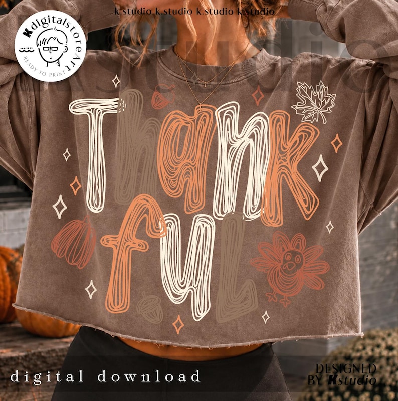 K&ouml;nnte beinhalten: Ein braunes bauchfreies Sweatshirt mit dem Wort Thankful in einer wei&szlig;en und orangefarbenen, krakeligen Schriftart. Das Design enth&auml;lt Strichzeichnungen eines Truthahns, einer Eichel, eines K&uuml;rbisses, eines Ahornblatts und kleiner Rauten.
