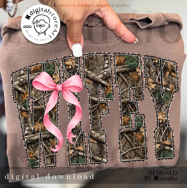 Puede incluir: Una sudadera marr&oacute;n con la palabra "WIFEY" en un patr&oacute;n de camuflaje, acentuada con un lazo rosa. El texto "digital download" est&aacute; en la parte inferior. Dise&ntilde;o de Kstudio.