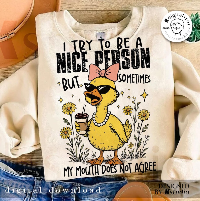 May include: A cream-coloured sweatshirt featuring a yellow duck wearing sunglasses, a pink bow, and a pearl necklace, holding a coffee cup. The text reads "I TRY TO BE A NICE PERSON BUT SOMETIMES MY MOUTH DOES NOT AGREE."