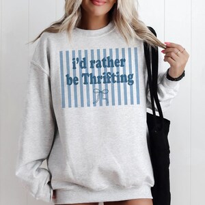 May include: A light gray sweatshirt with the text "I'd rather be Thrifting" in blue. The text is set against a background of blue vertical stripes and a bow graphic.