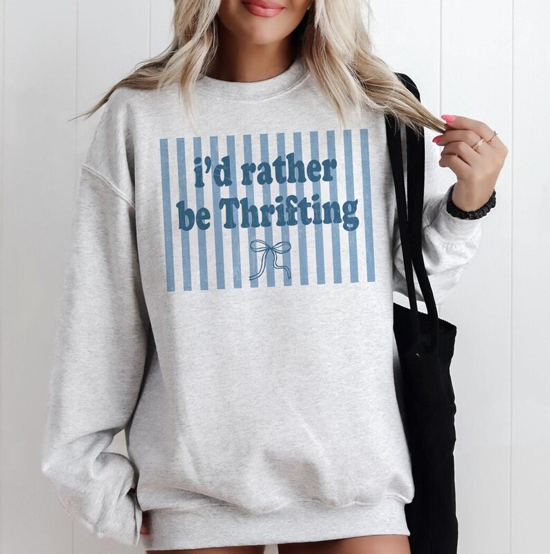 May include: A light gray sweatshirt with the text "I'd rather be Thrifting" in blue. The text is set against a background of blue vertical stripes and a bow graphic.