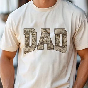 May include: Cream-colored t-shirt featuring the word "DAD" in a brown, textured font. The letters are filled with a detailed illustration of ducks in a marshy environment. The shirt is made of a soft material.