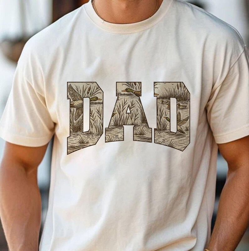 May include: Cream-colored t-shirt featuring the word "DAD" in a brown, textured font. The letters are filled with a detailed illustration of ducks in a marshy environment. The shirt is made of a soft material.