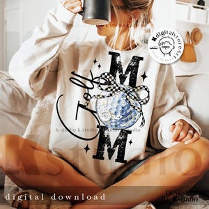 May include: A cream-colored sweatshirt with the word "MOM" in large black letters. A golf ball design is featured in the center, with a black and white checkered ribbon. The words "Golf Mom" are written in a script font.