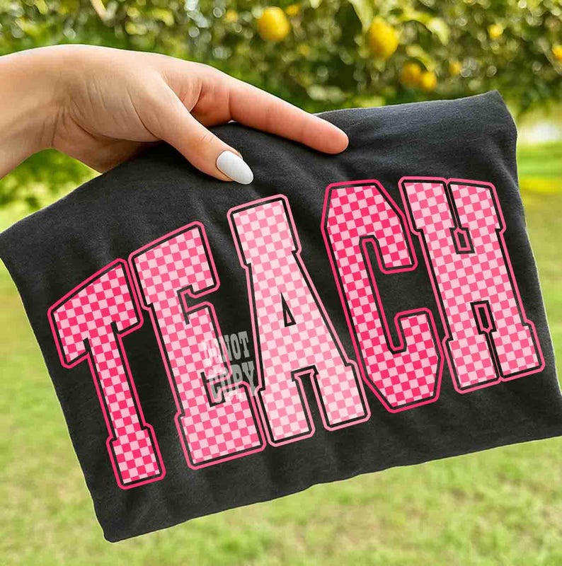 K&ouml;nnte beinhalten: Ein dunkelgraues T-Shirt mit dem Wort "TEACH" in gro&szlig;en, rosa karierten Buchstaben mit schwarzer Umrandung. Das T-Shirt wird von einer Hand vor einem verschwommenen gr&uuml;nen Hintergrund gehalten.