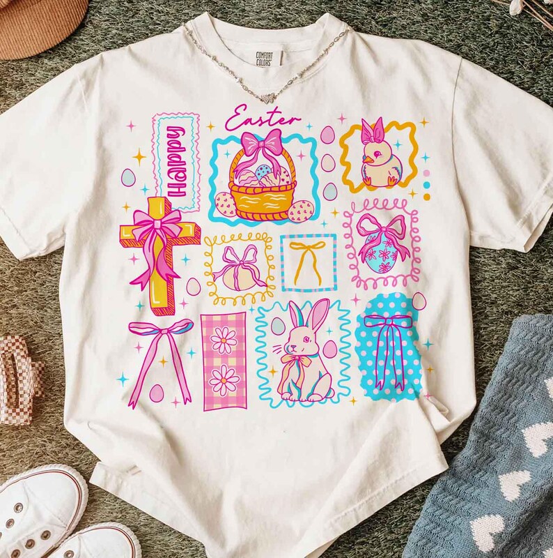 May include: White t-shirt with a colorful Easter-themed graphic. The design includes a cross, Easter basket, bunnies, eggs, and the word "Happy" and "Easter". The graphic is framed with pink, blue, and yellow borders.