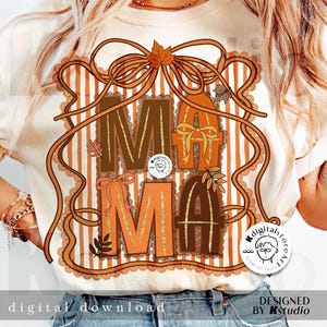 Op de afbeelding: Cr&egrave;mekleurig T-shirt met het woord "MAMA" in bruine en oranje blokletters, versierd met herfstaccenten. Het ontwerp bevat een strik, bladeren en een gestreepte achtergrond. De woorden "digital download" en "designed by Kstudio" zijn ook zichtbaar.