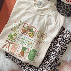 May include: Cream-colored t-shirt with a colorful Easter-themed graphic featuring a bunny, eggs, carrots, and teacups. Paired with leopard print pajama pants. The shirt has a Comfort Colors tag.