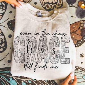 May include: Cream-colored sweatshirt with the text "even in the chaos GRACE still finds me." The word "GRACE" is filled with a floral design. The sweatshirt is folded and held by hands.