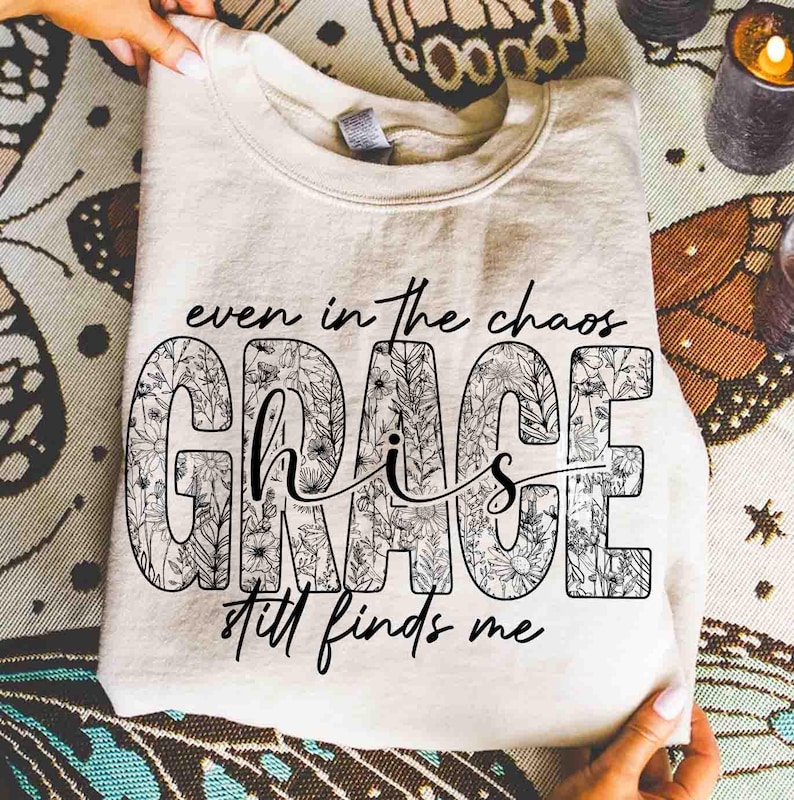 May include: Cream-colored sweatshirt with the text "even in the chaos GRACE still finds me." The word "GRACE" is filled with a floral design. The sweatshirt is folded and held by hands.