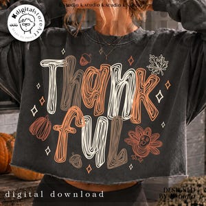 K&ouml;nnte beinhalten: Ein anthrazitfarbenes, bauchfreies Sweatshirt mit dem Wort Thankful in einer wei&szlig;en, orangefarbenen und braunen Kritzel-Schriftart. Das Design enth&auml;lt Illustrationen eines Truthahns, von Eicheln, einem Ahornblatt und kleinen Rauten.