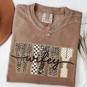 May include: A brown Comfort Colors t-shirt with the word "wifey" in a decorative font. The letters feature leopard print, checkered, and polka dot patterns. A silver necklace with a pendant rests on the shirt.