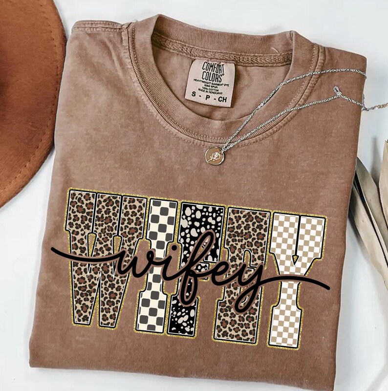 May include: A brown Comfort Colors t-shirt with the word "wifey" in a decorative font. The letters feature leopard print, checkered, and polka dot patterns. A silver necklace with a pendant rests on the shirt.