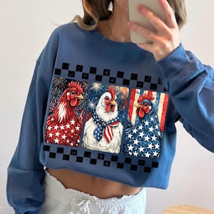 May include: Blue cropped sweatshirt featuring three chickens with patriotic designs. The chickens are depicted with red, white, and blue colors, including stars and stripes, and fireworks. A black and white checkered pattern borders the chicken images.