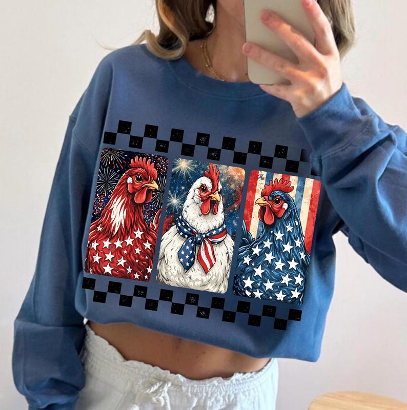 May include: Blue cropped sweatshirt featuring three chickens with patriotic designs. The chickens are depicted with red, white, and blue colors, including stars and stripes, and fireworks. A black and white checkered pattern borders the chicken images.
