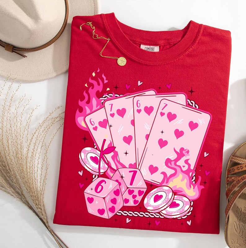 May include: Red crew neck t-shirt featuring a graphic of playing cards, dice, and poker chips with pink hearts. The design includes flames and the numbers 6 and 7. A gold necklace and a beige hat are visible.