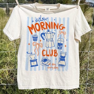 May include: Cream-colored t-shirt with a coffee-themed graphic. The design features the words "Slow Morning Club" in orange, along with blue and orange illustrations of coffee-related items like a coffee maker, grinder, and cup. The shirt is hanging on a clothesline.