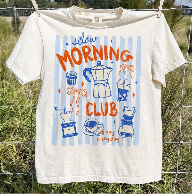 May include: Cream-colored t-shirt with a coffee-themed graphic. The design features the words "Slow Morning Club" in orange, along with blue and orange illustrations of coffee-related items like a coffee maker, grinder, and cup. The shirt is hanging on a clothesline.