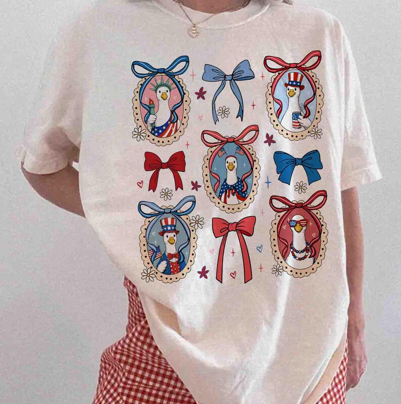 Op de afbeelding: Cr&egrave;mekleurig T-shirt met een patriottisch ontwerp. Het shirt toont negen ovale kaders met ganzenillustraties in patriottische kleding, omringd door strikken, harten en bloemaccenten. Het ontwerp bevat rood, wit en blauw.