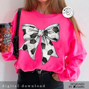 May include: A pink sweatshirt with a black and white soccer ball bow design. The sweatshirt has a crew neck and long sleeves.