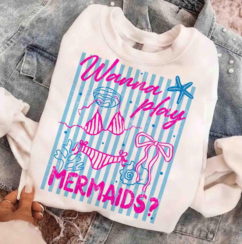 May include: White sweatshirt with a graphic design featuring the text "Wanna play MERMAIDS?" in pink, along with blue and pink illustrations of swimwear and seashells. The design is set against a background of blue and white vertical stripes.